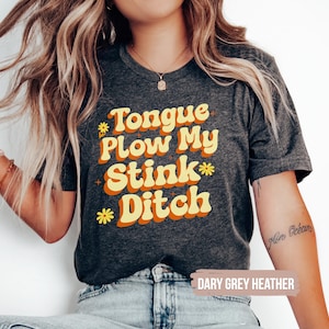May include: Dark grey heather t-shirt with the text "Tongue Plow My Stink Ditch" in orange and yellow retro font. The shirt has a crew neck and short sleeves. The text "DARY GREY HEATHER" is at the bottom.