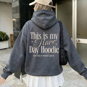 May include: Dark heather gray hoodie with the text "This is my Flare Day Hoodie" in a decorative font. The hoodie also has the text "Operating at minimal capacity" below the main text. The hoodie is part of a casual wear collection.