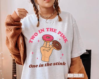 May include: Ash grey t-shirt featuring the text "TWO IN THE PINK" above a cartoon hand holding two donuts, one pink and one brown. The text "One in the stink" is below the hand.