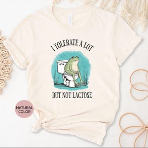 May include: A white t-shirt with the text "I tolerate a lot but not lactose" and a cartoon frog sitting on a toilet.