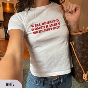 May include: White t-shirt with the red text "WELL BEHAVED WOMEN RARELY MAKE HISTORY." The shirt is paired with blue jeans. A brown handbag with a monogram pattern is visible.