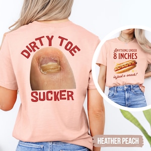 May include: A heather peach t-shirt featuring a large photographic print of a toe on the back with the red text DIRTY TOE SUCKER. The front displays a hot dog graphic with the text Anything under 8 INCHES is just a snack.