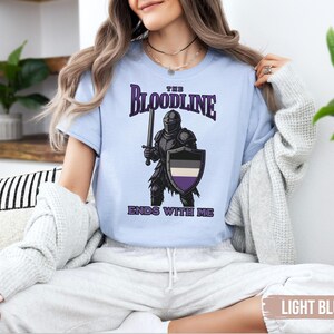 Puede incluir: Camiseta azul claro con un gr&aacute;fico de un caballero sosteniendo una espada y un escudo. El escudo tiene los colores de la bandera del orgullo asexual. El texto en la camiseta dice "The Bloodline Ends With Me."