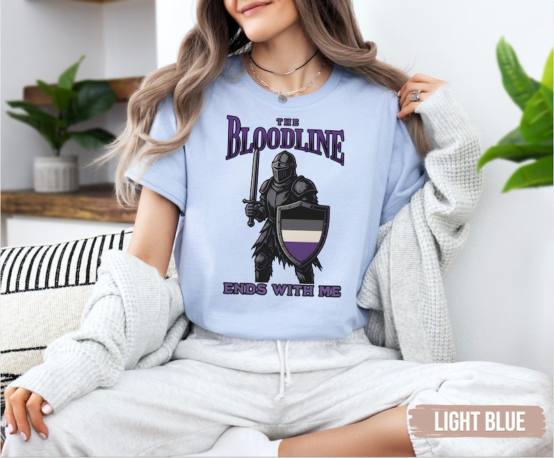 Puede incluir: Camiseta azul claro con un gr&aacute;fico de un caballero sosteniendo una espada y un escudo. El escudo tiene los colores de la bandera del orgullo asexual. El texto en la camiseta dice "The Bloodline Ends With Me."