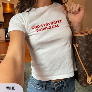 May include: White t-shirt with the red text "GOD'S FAVORITE PANSEXUAL". The shirt is a crew neck style and is paired with blue jeans. A brown handbag is visible in the background.