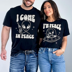 May include: Two black t-shirts with white graphics. One says "I COME IN PEACE" with an alien holding a ray gun. The other says "I'M PEACE" with an alien in a UFO. Both shirts feature a retro font.