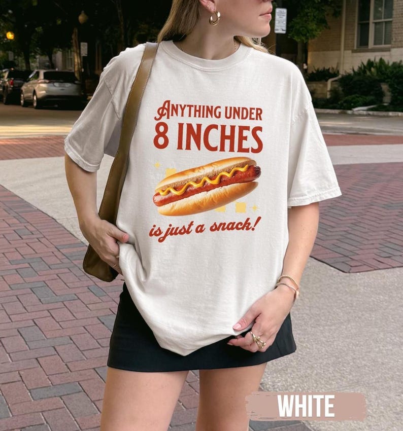 May include: Cream-colored t-shirt with the text "Anything Under 8 Inches is just a snack!" and a graphic of a hot dog. The hot dog has mustard and is on a bun. The shirt is a casual style.