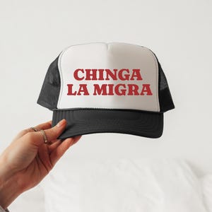 Può includere: Cappellino da camionista nero e bianco con la scritta "CHINGA LA MIGRA" in lettere rosse sul davanti. Il cappellino ha una visiera nera e un retro in rete.