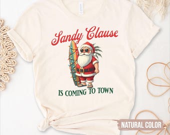 May include: Cream-coloured t-shirt with the text "Sandy Clause Is Coming To Town." The graphic features Santa Claus in sunglasses, holding a surfboard decorated with lights. The shirt is a natural colour.