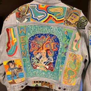 May include: A light blue denim jacket with colorful painted designs. The jacket features a large painted image of a woman with red hair and an octopus, with the text "She kept her children on Buckingham Green". Other designs include a pirate, a shark, a fish, and a cartoon character.
