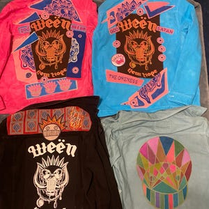 May include: Four denim jackets with custom patches and designs. The jackets are in various colors, including pink, blue, black, and light blue. The patches feature the band Ween's logo, the words "God", "Satan", "The Oneness", and "New Hope", and other designs. The jackets are all handmade and available in all sizes.