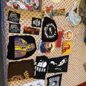 May include: A collection of black t-shirts with various graphic designs, including a skull with a microphone, a person with a gun, and the text "It's Gonna Be A Long Night". There are also several patches and stickers with various designs.