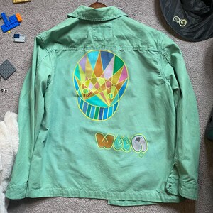 May include: A light green jacket with a colorful graphic on the back. The graphic features a geometric design with orange, yellow, blue, and green colors. The word "Woog" is printed below the graphic.