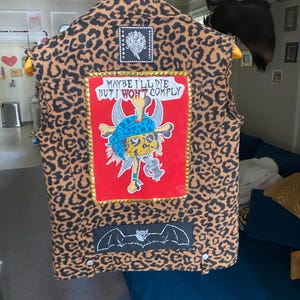 May include: A brown and black leopard print vest with patches. The patches include a skull and crossbones with the text "Maybe I'll Die But I Won't Comply", a bat, and a black and white image of a face.