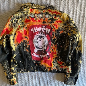 May include: Black denim jacket with a distressed, splattered design in yellow and red. The back features a large patch with the word "Ween" and a graphic. The collar is adorned with gold spikes.