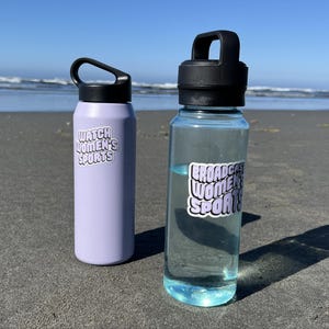 May include: Two water bottles with stickers that say "Watch Women's Sports" and "Broadcast Women's Sports" on a sandy beach.