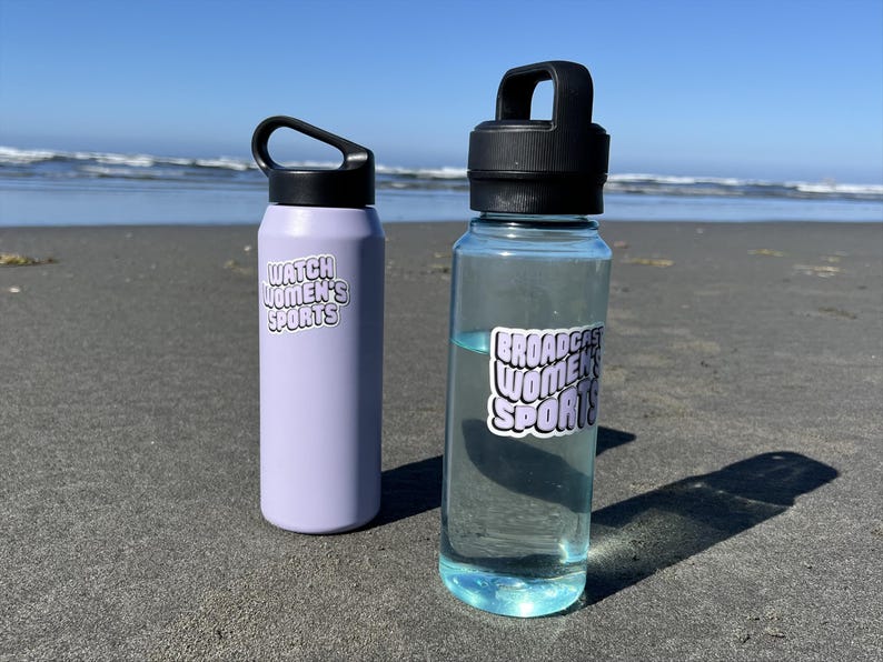 May include: Two water bottles with stickers that say "Watch Women's Sports" and "Broadcast Women's Sports" on a sandy beach.