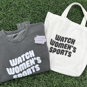 May include: A grey sweatshirt with the text "WATCH WOMEN'S SPORTS" in white lettering. A white tote bag with the same text in black lettering. Both items are on a green grassy surface.