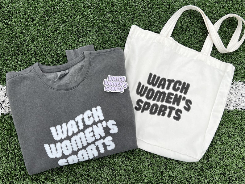 May include: A grey sweatshirt with the text "WATCH WOMEN'S SPORTS" in white lettering. A white tote bag with the same text in black lettering. Both items are on a green grassy surface.