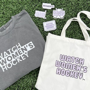 May include: A gray sweatshirt and a white tote bag both feature the text "Watch Women's Hockey" in different styles. The tote bag has a purple outline around the text and a hockey stick graphic. There are several stickers with the same text in different colors and styles.