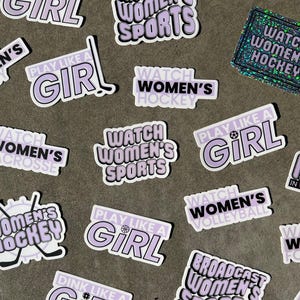 May include: A collection of stickers with various designs and text, including "Watch Women's Sports", "Play Like a Girl", and "Watch Women's Hockey". The stickers are purple and white with black text.