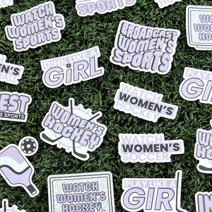 May include: A collection of stickers with various slogans and designs related to women's sports. The stickers are purple and white with text such as "Watch Women's Hockey", "Invest in Women's Sports", and "Play Like a Girl".