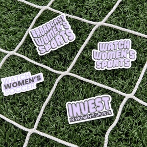 May include: Four stickers with purple text on a white background. The stickers say "Broadcast Women's Sports", "Watch Women's Soccer", "Watch Women's Sports", and "Invest in Women's Sports". The stickers are on a green artificial grass surface with a white net.
