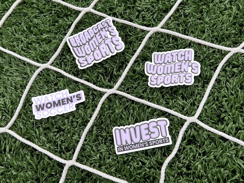 May include: Four stickers with purple text on a white background. The stickers say "Broadcast Women's Sports", "Watch Women's Soccer", "Watch Women's Sports", and "Invest in Women's Sports". The stickers are on a green artificial grass surface with a white net.