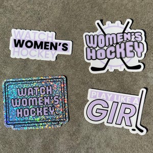 May include: Four stickers with a purple and white color scheme. The stickers feature the text "Watch Women's Hockey" and "Play Like A Girl!" with a hockey stick graphic. One sticker has a glittery background.