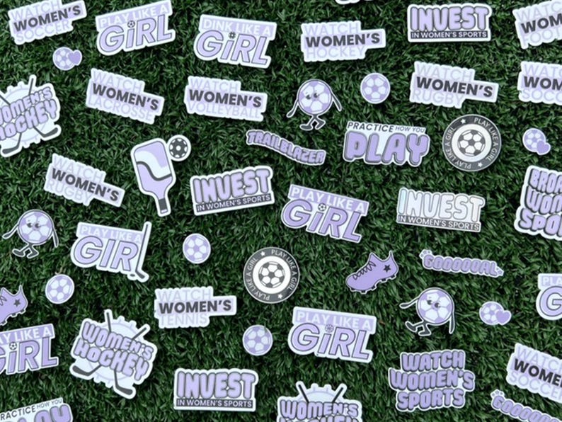 May include: A collection of purple and white stickers with various sports-themed designs. The stickers feature phrases like "Play Like a Girl", "Watch Women's Soccer", and "Invest in Women's Sports".