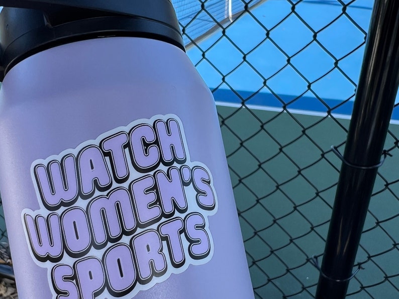 May include: A lavender water bottle with a sticker that says "Watch Women's Sports" in a retro font.