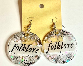 May include: A pair of silver glitter earrings with a clear resin base. The earrings are round and have the word "folklore" written on them in black.