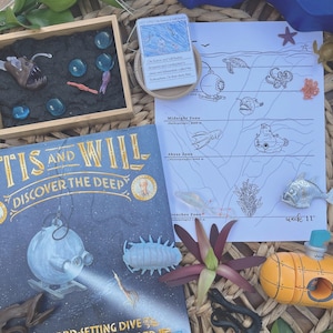 May include: A book titled "The Record-Setting Dive of the Bathysphere" by Barb Roe, featuring illustrations of a submarine and a deep-sea creature. The book is surrounded by various toys, including a miniature submarine, a sea creature, and a small wooden box with blue and green glass beads.