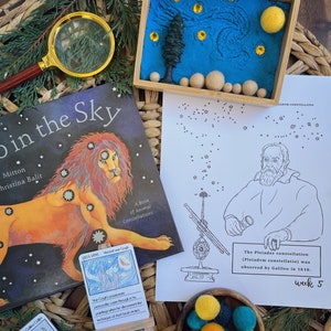 May include: A children's book titled "Join the Sky: A Book of Animal Constellations" with a colorful illustration of a lion on the cover. A small easel holds a card with a painting of "Starry Night" by Vincent van Gogh. A black and white worksheet with a drawing of Galileo Galilei and a telescope. The Pleiades constellation is labeled on the worksheet. The text "The Pleiades constellation (Pleiadvm constellatio) was observed by Galileo in 1610. Week 5" is printed on the worksheet. A small wooden box with a blue sand tray and yellow felt stars and a yellow felt sun. A bowl of yellow, teal, and black felt balls.