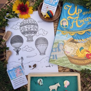 Könnte beinhalten: Eine Auswahl an Lernmaterialien, darunter eine Malvorlage mit Heißluftballons, ein Buch mit dem Titel "Up and Away" und verschiedene Spielfiguren. Ein Holztablett enthält Miniatur-Heißluftballons, eine Ente, ein Schaf und ein Huhn. Eine Blume und Grünzeug umgeben die Gegenstände.