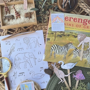 Könnte beinhalten: Eine Nahaufnahme eines pädagogischen Aktivitätensatzes für Kinder mit einer Malvorlage mit Noten und Tieren, einer Holzkiste mit Tierfiguren und Reis, einem Buch über das Leben eines Komponisten und einem Satz Papierpuppen mit Stöcken. Der Buchumschlag zeigt ein farbenfrohes Zirkuszelt und den Text "1835-1921 Camille Saint-Saëns". Die Malvorlage zeigt einen Löwen, einen Elefanten, einen Schwan und einen Esel. Die Puppen umfassen ein Zebra, ein Känguru, einen Schwan und einen Fisch.