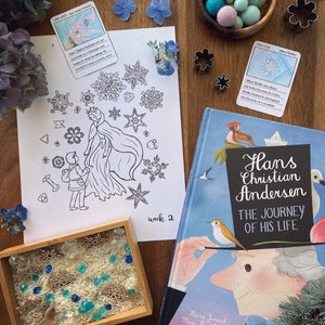 May include: A wooden table displays a children's book, coloring page, and sensory bin. The book cover reads "Hans Christian Andersen: The Journey of His Life." The coloring page features a princess and snowflakes. The bin contains rice, wooden snowflakes, and blue gems.