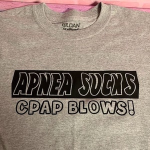 May include: Gray t-shirt with black text that reads "APNEA SUCKS CPAP BLOWS!" The text is in a bold, outlined font.