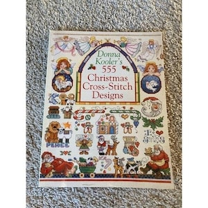 May include: A softcover book titled Donna Kooler's 555 Christmas Cross-Stitch Designs. The cover features a grid of colorful cross-stitch patterns including angels, Santa Claus, a gingerbread house, candy canes, and various holiday motifs.
