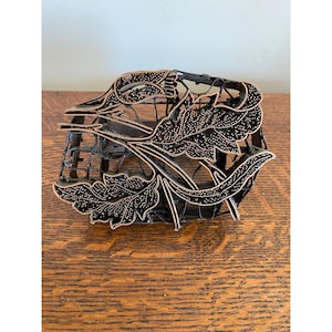 May include: A copper batik tjap stamp featuring a detailed floral and leaf design with intricate wirework patterns. The metal tool has a dark, oxidized finish on the frame and a polished copper surface on the raised stamping elements.