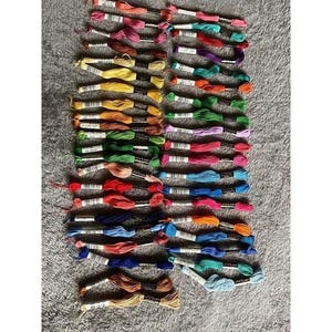 May include: A collection of embroidery floss skeins arranged in two vertical columns. The threads feature a wide spectrum of colors including shades of red, blue, green, yellow, orange, purple, pink, brown, and white, each with a paper label.
