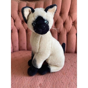 May include: A plush Siamese cat toy featuring cream-colored fur on the body, dark brown fur on the ears, face, and paws, and bright blue eyes. The stuffed animal is in a seated position with pointed ears and long white whiskers on each side.