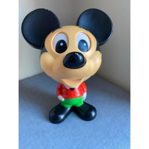 May include: A plastic Mickey Mouse figurine featuring a large head with black ears, a tan face, and white eyes with black pupils. The character wears a red long-sleeved shirt, green pants, and black shoes. The figure has a small black nose.
