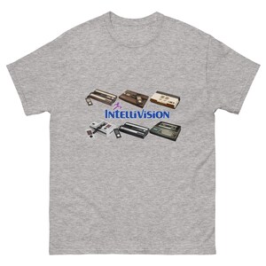 May include: A heather gray t-shirt featuring a collection of vintage Intellivision game consoles. The consoles are in various colors, including brown, black, and white. The Intellivision logo is displayed in blue and purple.
