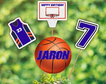 May include: A basketball themed centerpiece with a basketball, a basketball jersey, a basketball hoop, and the number 7. The centerpiece also includes a wooden box with succulents and the name "Jaron" written on the basketball.