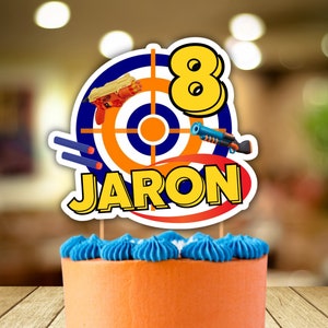 Könnte beinhalten: Ein orangefarbener Kuchen mit blauer Glasur und Streuseln. Ein Tortenaufsatz zeigt ein Zielscheibendesign mit Spielzeugpistolen, die Zahl 8 und den Namen "JARON". Der Kuchen steht auf einem blauen Ständer, ideal für einen Geburtstag.