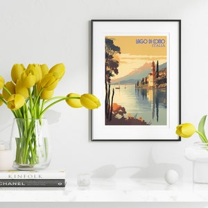 May include: A framed vintage travel poster with a scenic view of Lake Como in Italy. The poster features a lake with a church building on a peninsula, mountains in the background, and a sailboat on the water. The text on the poster reads "Lago di Como, Italia".