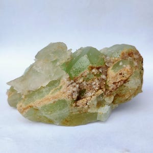 May include: A cluster of raw, uncut green crystals with a translucent appearance. The crystals are clustered together with a light brown matrix and a white background. The crystals have a natural, rough texture.