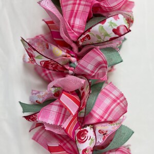 May include: A garland composed of layered pink ribbons in various patterns, including plaid, floral, and striped designs. The ribbons are in shades of pink and red, with accents of green. The garland is likely used for decoration.