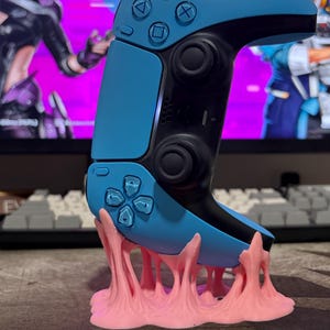 May include: A blue and black video game controller is displayed on a pink, bubblegum-like stand. The controller has buttons with symbols and a directional pad. The stand appears to be made of a soft, textured material.